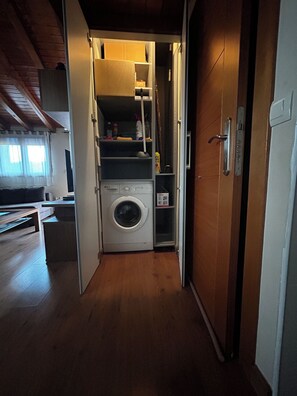 A laundry room with a washing machine, shelves with cleaning supplies, and a wooden floor. - Stargazing Apartment with Terrace & Telescope in a Historic Sierra Village (San Martín del Castañar)