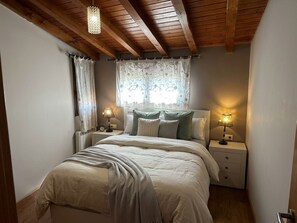 A bedroom with a wooden ceiling, a bed with a white comforter, two bedside lamps, and a window with a patterned curtain.