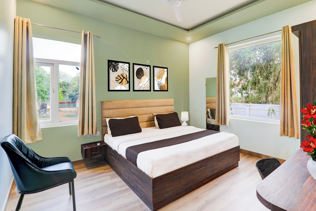 A hotel room with a bed, a chair, a desk, and a window with curtains. There are three framed pictures on the wall.