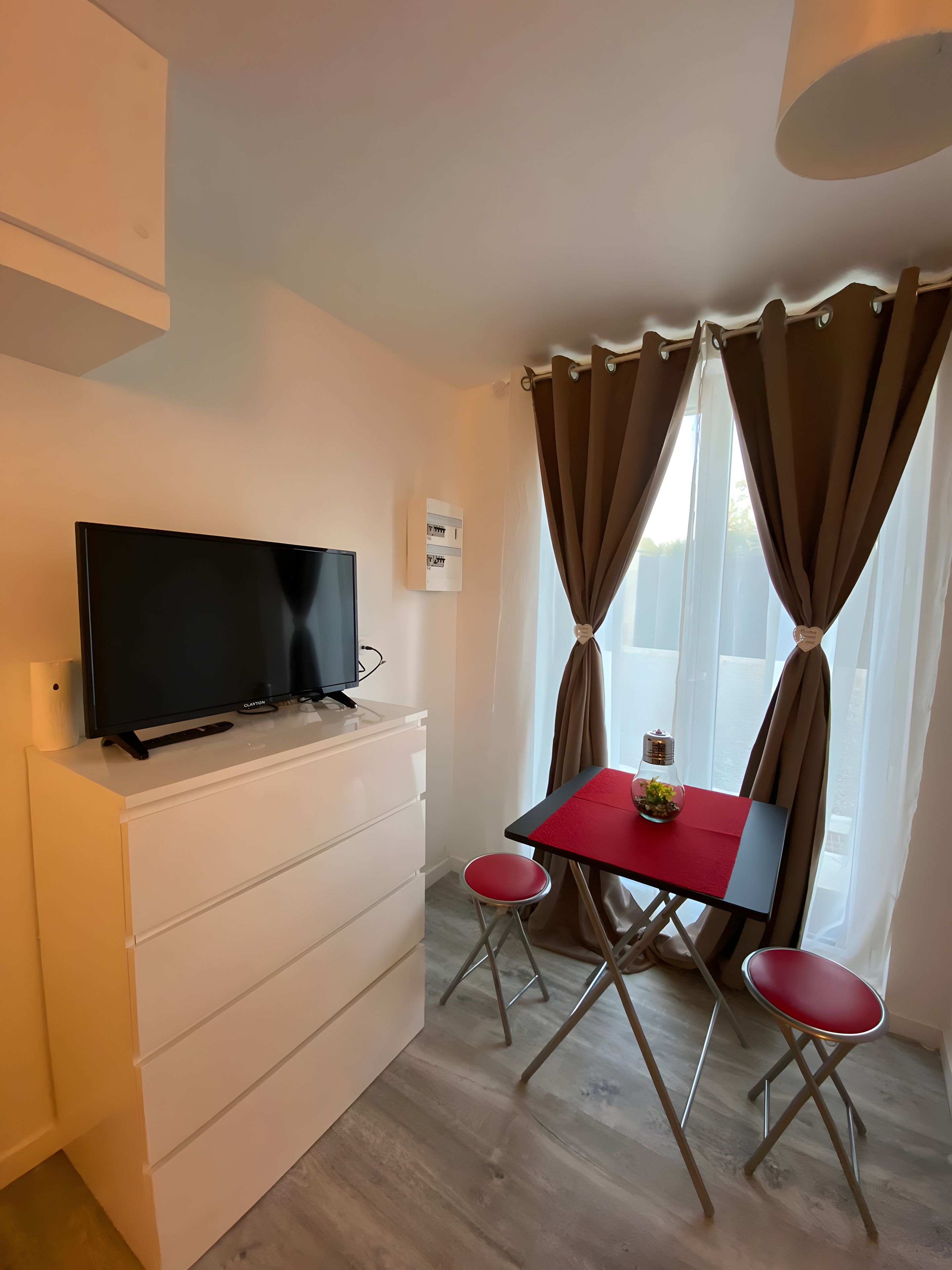 A room with a flat-screen TV on a white dresser, a small table with a red top, two red chairs, and a window with curtains.