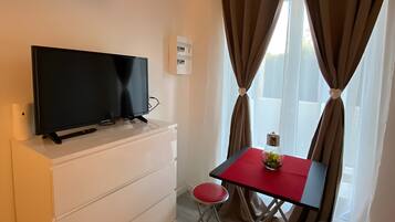A room with a flat-screen TV on a white dresser, a small table with a red top, two red chairs, and a window with curtains.