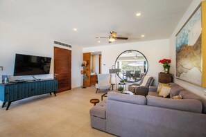 Ein modernes Wohnzimmer mit großem Flachbildfernseher, einer grauen Couch, einem Holzkasten und einem Deckenventilator.
