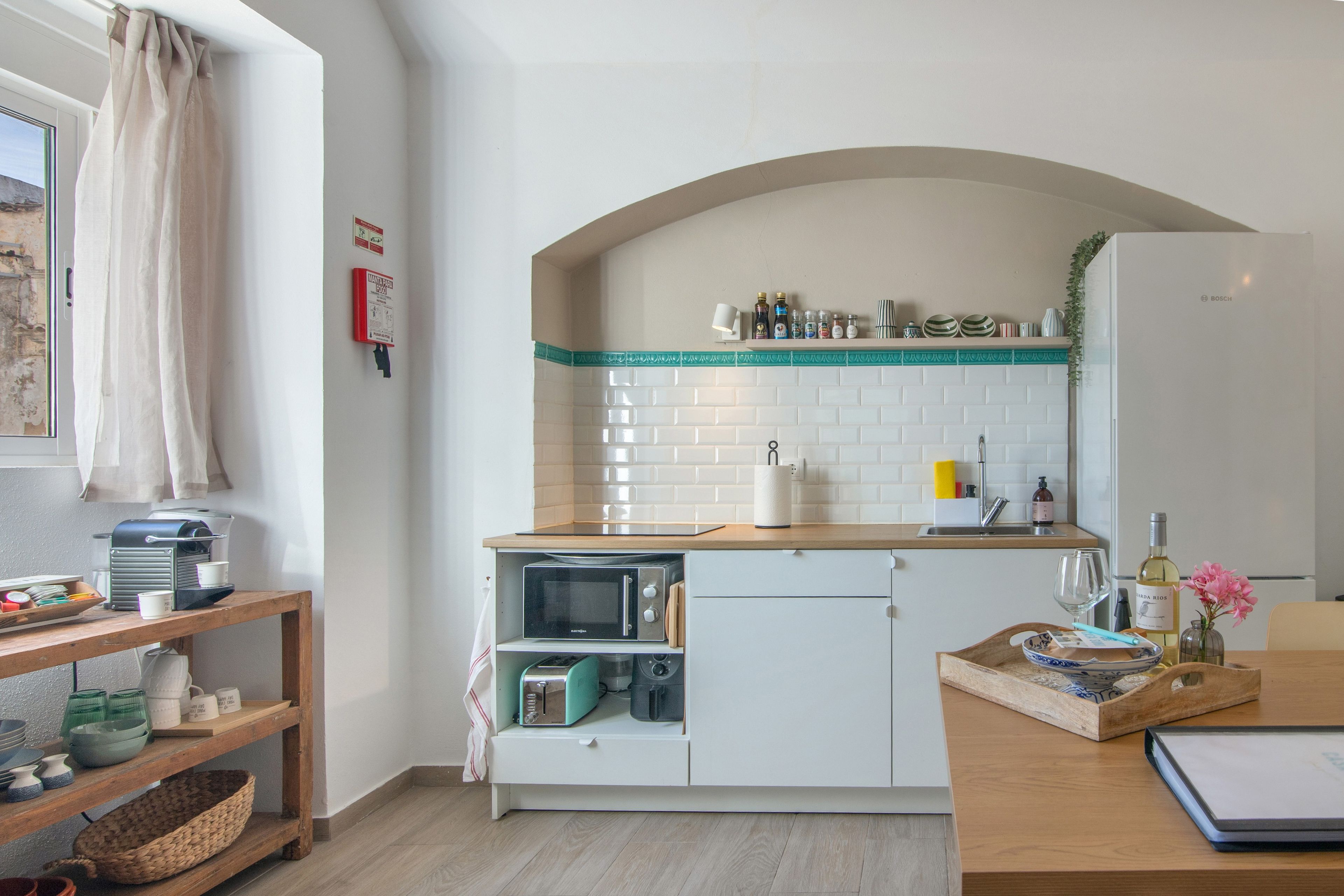 Una cucina compatta con forno a microonde, tostapane e lavello. Si vede uno scaffale in legno con stoviglie e una finestra con tende.