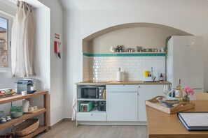 A compact kitchen with a microwave, toaster, and a sink. There is a wooden shelf with kitchenware and a window with curtains.