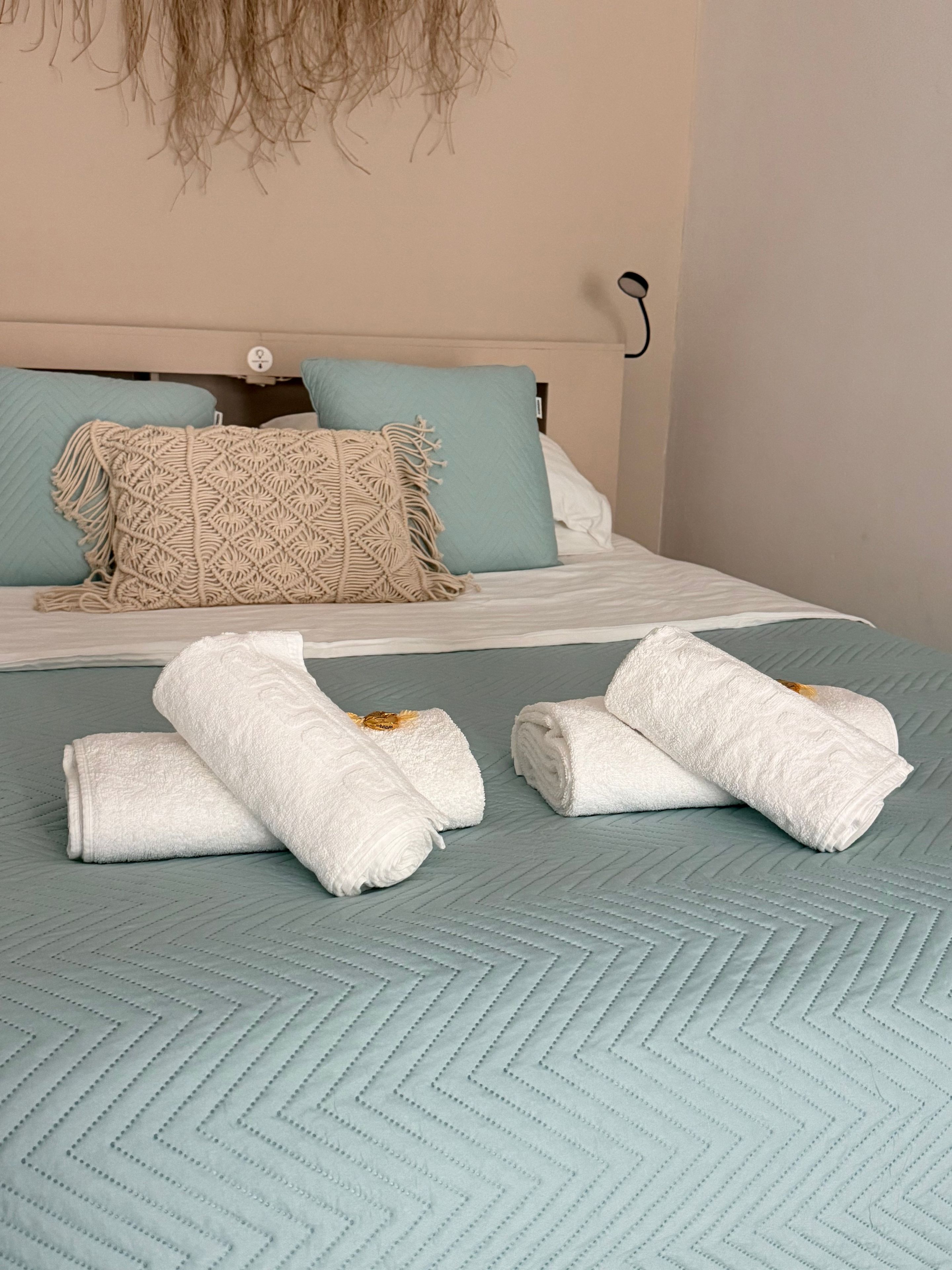 A bed with a light blue quilt, two pillows, and a macrame throw. There are three rolled white towels on the bed.