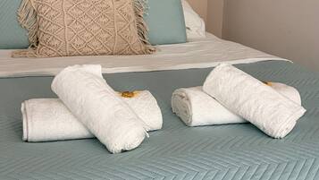 A bed with a light blue quilt, two pillows, and a macrame throw. There are three rolled white towels on the bed.