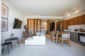 A modern living room with a sofa, dining table, and chairs, a kitchen with wooden cabinets, and a hallway leading to a bedroom. - Paraíso MarAlma 2BR Rooftop Vista al Mar Bucerías (Bucerías)