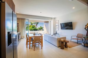 A modern living room with a dining area, a sofa, a TV mounted on the wall, and a view of greenery outside. - Paraíso MarAlma 2BR Rooftop Vista al Mar Bucerías (Bucerías)