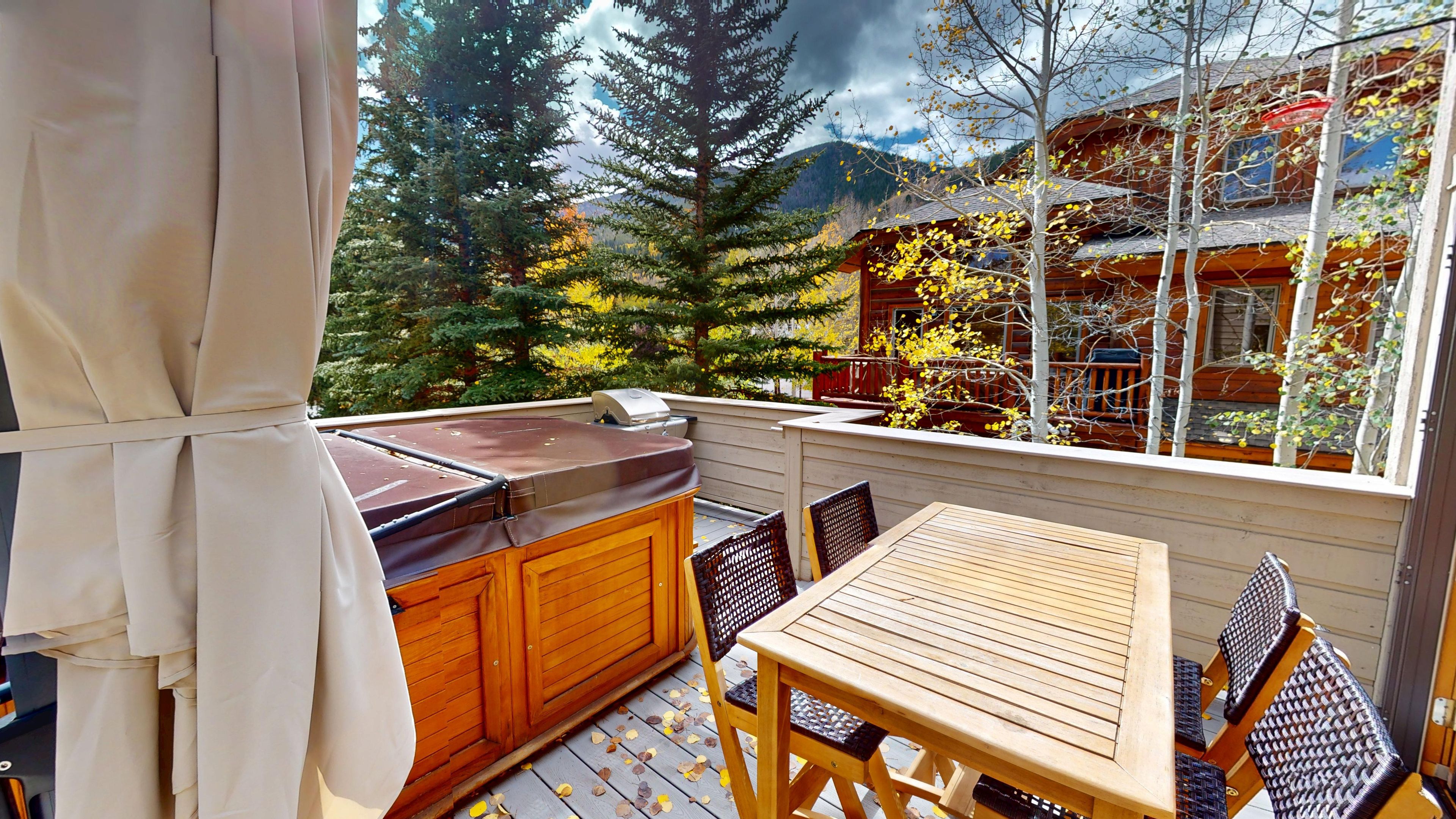 A balcony with a wooden table and chairs, a barbecue grill, and a view of a cabin surrounded by trees.