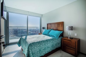 A bedroom with a large bed, a nightstand, a lamp, and a view of a cityscape through a large window. - Ocean views in luxury complex. (Nuevo Vallarta)