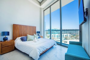 A hotel room with a large bed, a wooden headboard, a nightstand with a lamp, a blue chair, and a view of a building through large windows. - Best roof top Pool in Puerto Vallarta - 3 Bedroom (Nuevo Vallarta)