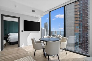 A modern hotel room with a large window, a flat-screen TV, a bed, a dining area with chairs, and a view of the cityscape. - 30th Floor Luxury Suite with Bay Views & Rooftop Pool (Miami)