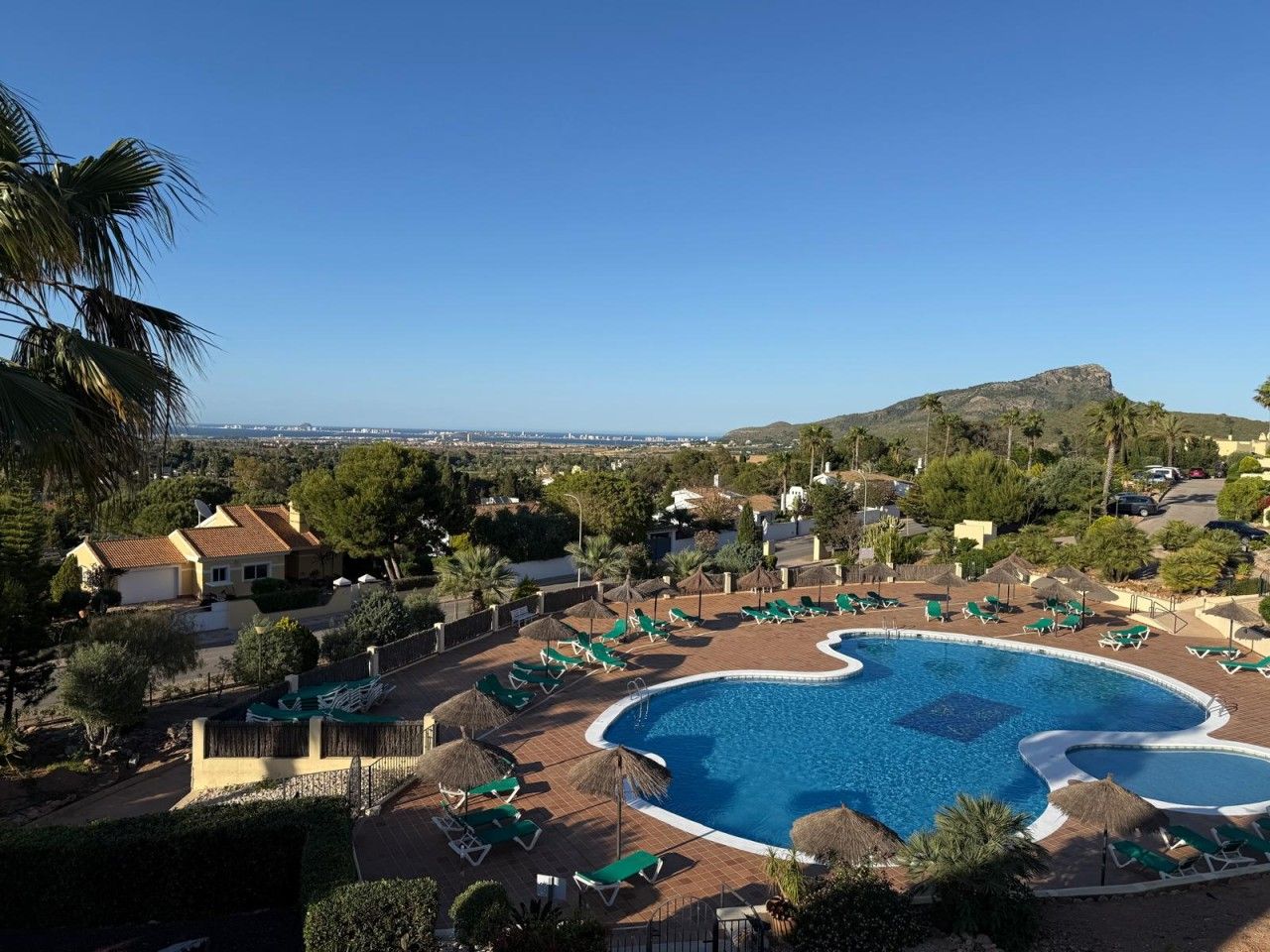 A resort with a large swimming pool surrounded by lounge chairs and umbrellas, set against a backdrop of hills and clear skies.