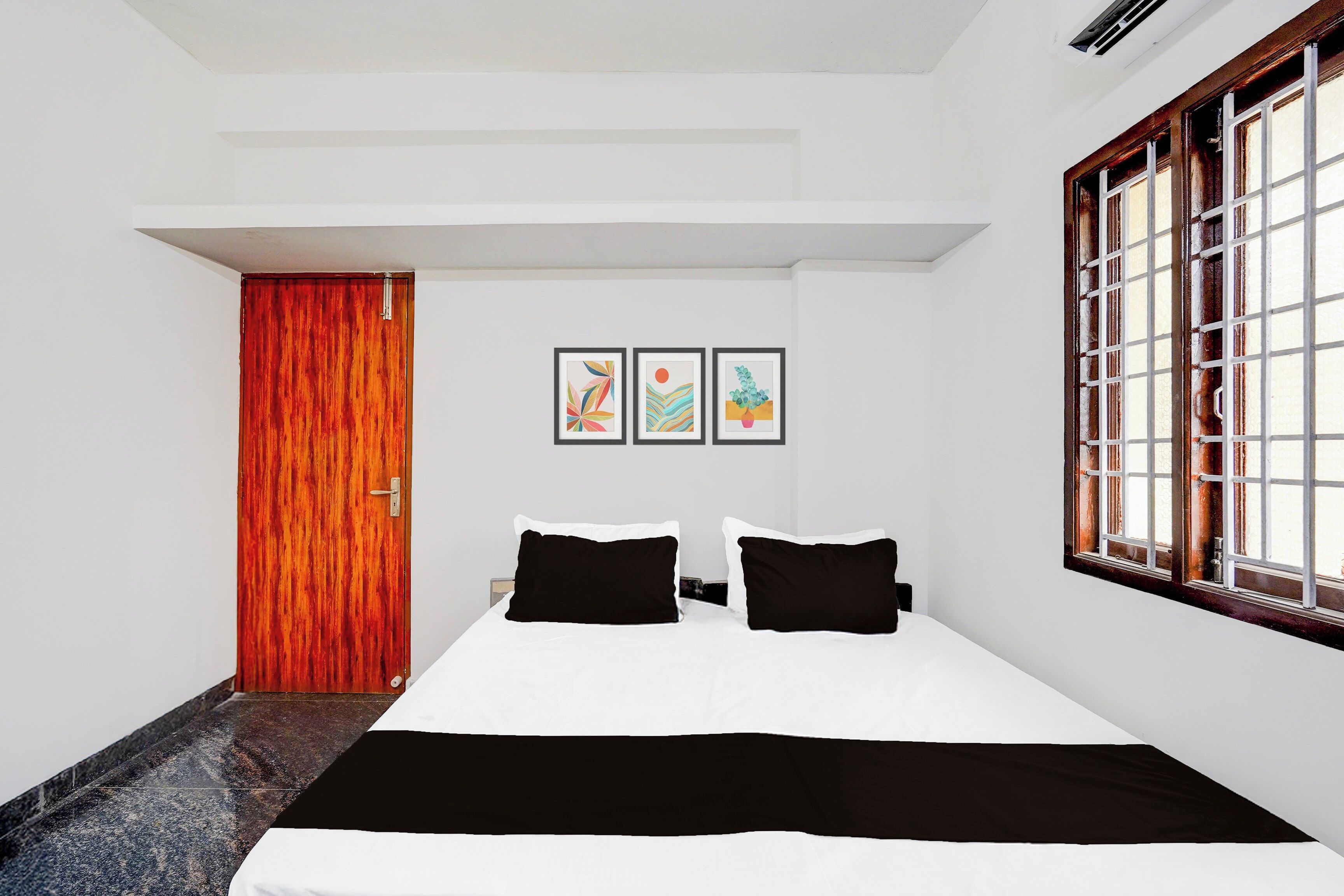 A bedroom with a bed, a window with bars, a door, and three framed pictures on the wall.