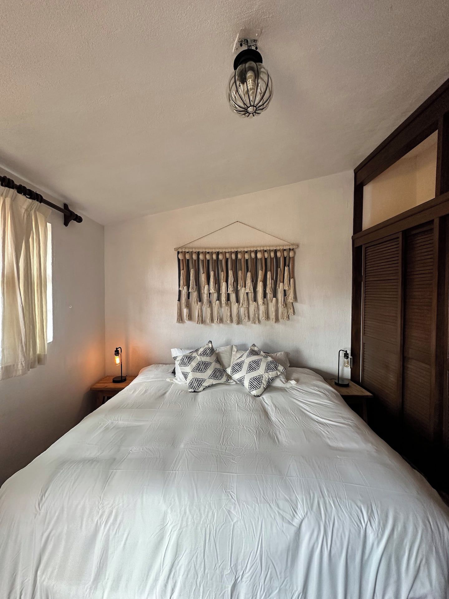 A bedroom with a bed, a wall-mounted macrame piece, a bedside lamp, and a hanging light fixture.