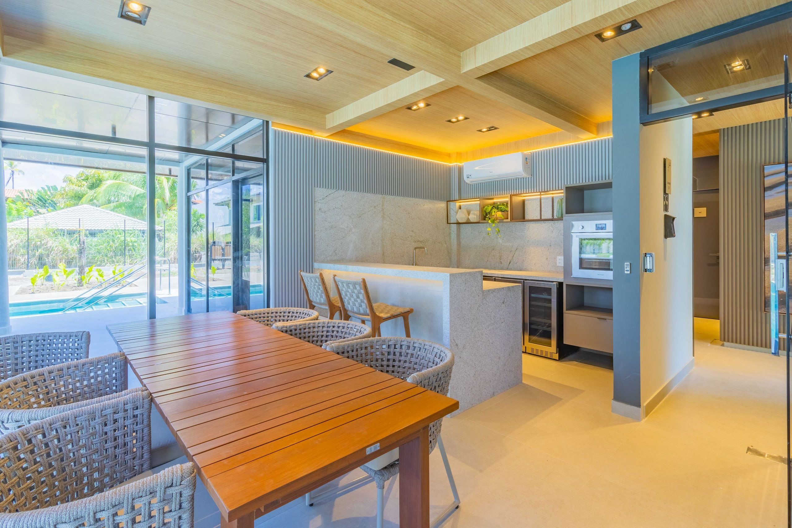 A modern kitchen with a central island, a dining area with wicker chairs, and large windows offering a view of a pool and greenery.