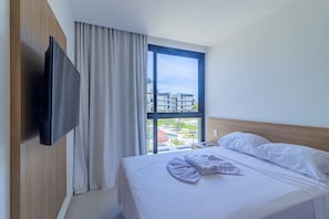 Habitación de hotel con una cama grande, un televisor en la pared y un ventanal con vistas a edificios y zonas verdes.