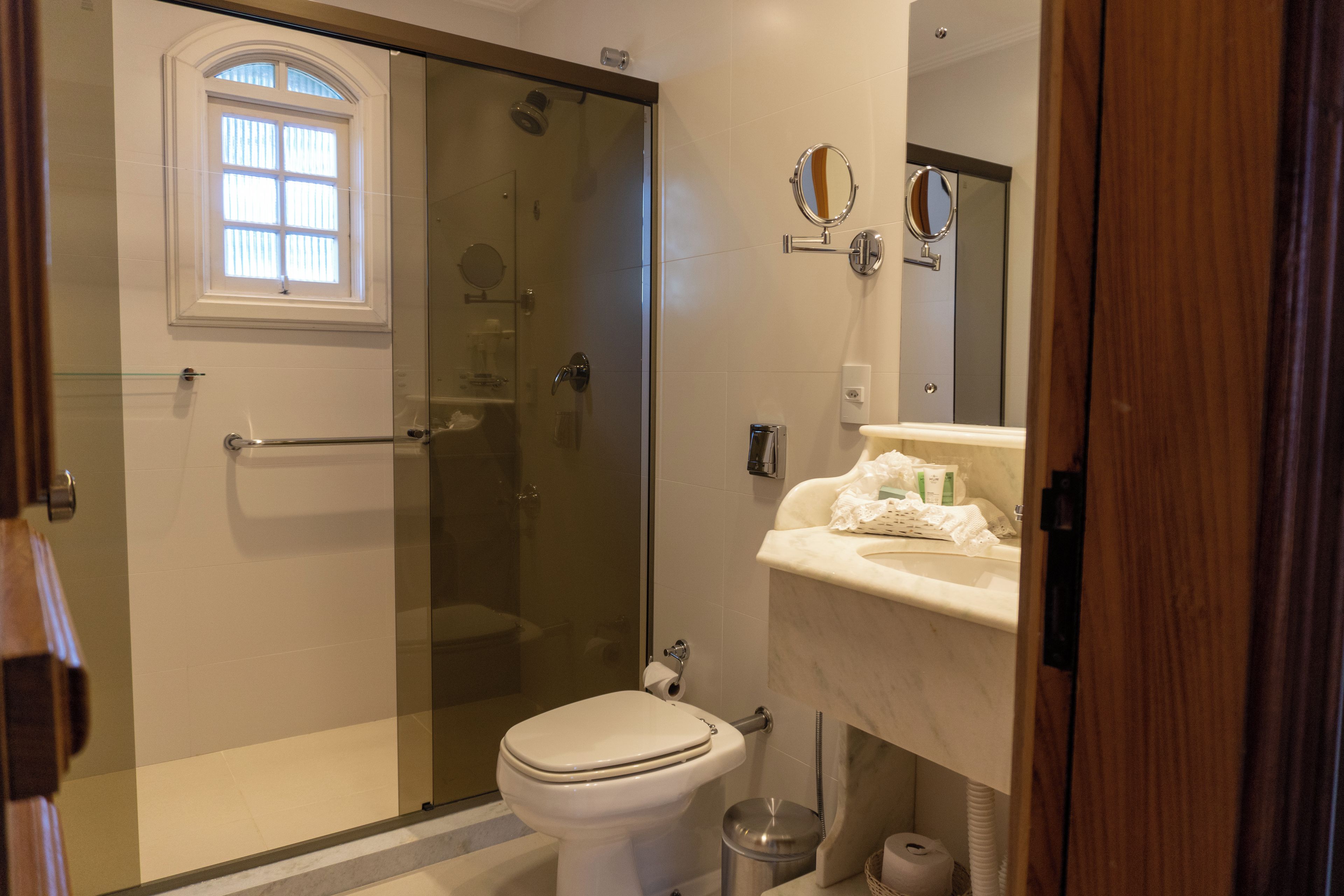 Un baño con ducha envuelta en vidrio, inodoro, lavabo con espejo y ventana sobre el inodoro.