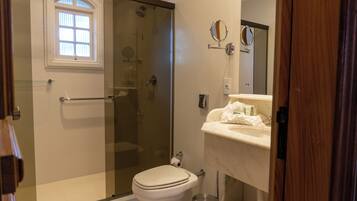 Un baño con ducha envuelta en vidrio, inodoro, lavabo con espejo y ventana sobre el inodoro.