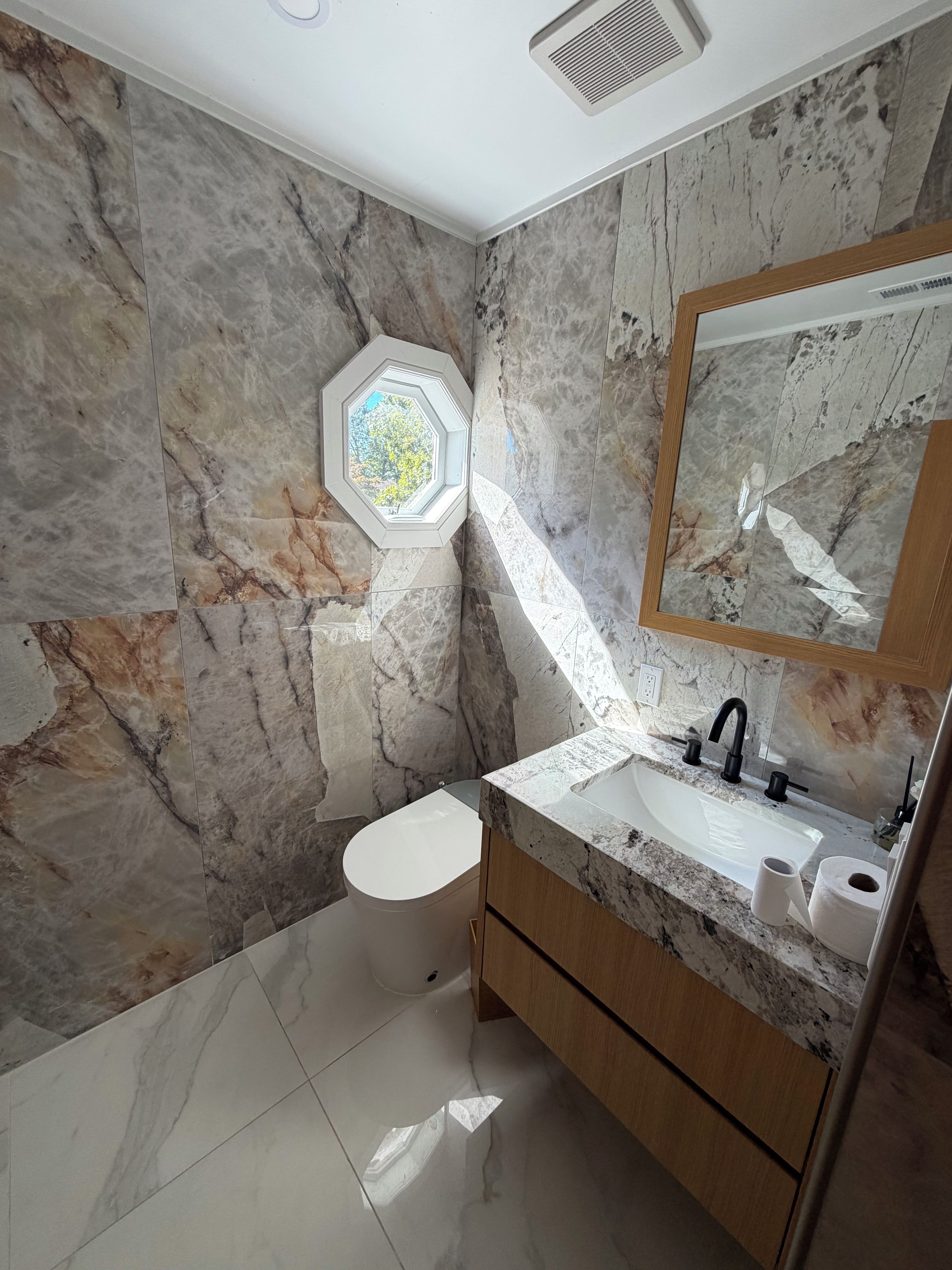 A bathroom with a marble wall, a hexagonal skylight, a white toilet, a sink with a mirror, and wooden cabinets.