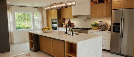 A modern kitchen with a central island, wooden cabinets, stainless steel appliances, and a large window.