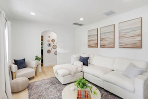A modern living room with a white sofa, a coffee table, and a rug. There are three wall-mounted paintings and a door leading to another room.