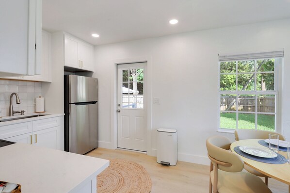 A modern kitchen with white cabinetry, a stainless steel refrigerator, a dining area with a table and chairs, and a window with a view of greenery.