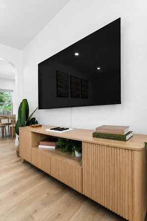A modern living room with a large flat-screen TV mounted on a white wall, a wooden console table with books, and a potted plant.