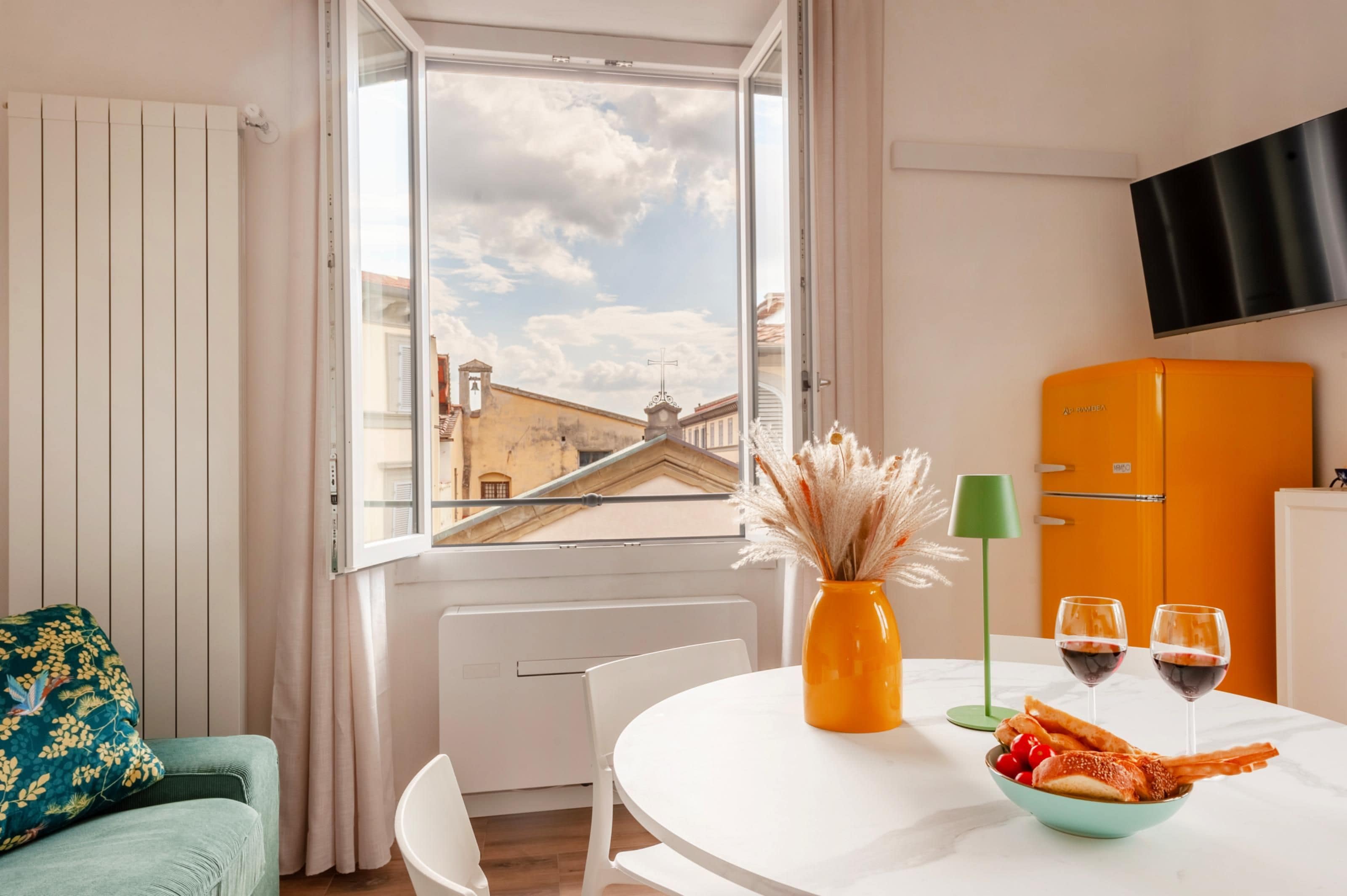 Une pièce comprenant une table à manger, un canapé, un téléviseur fixé au mur et une fenêtre donnant sur des immeubles.