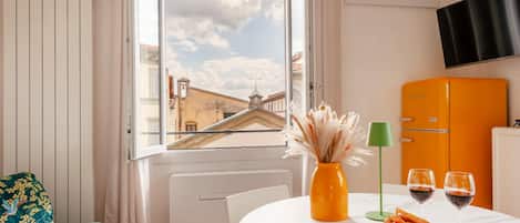 A room with a dining table, a sofa, a TV mounted on the wall, and a window with a view of buildings.