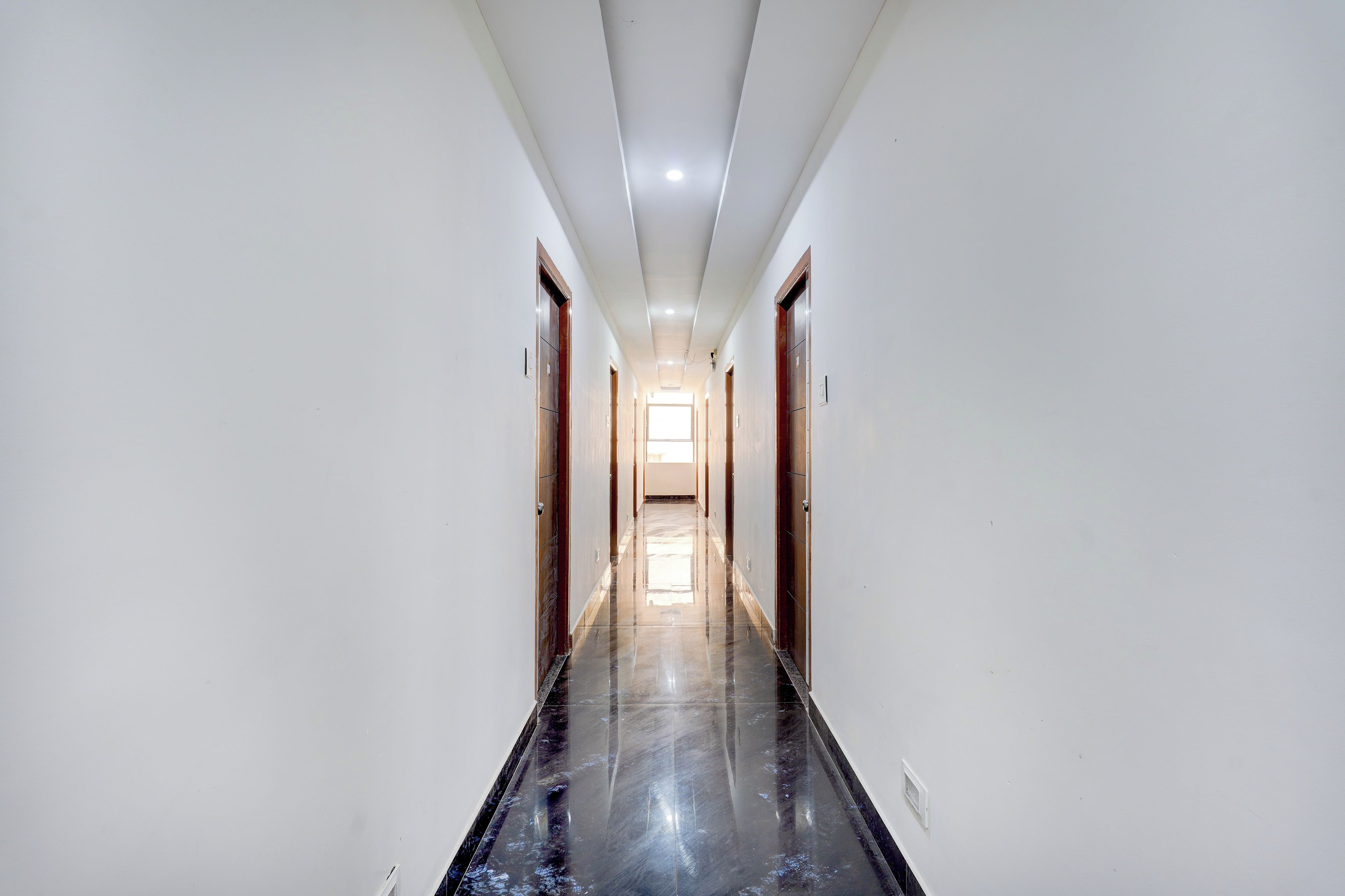 A long, narrow hallway with white walls and a shiny black floor, leading to a bright room at the end.