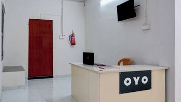 A reception desk with a sign reading 'OYO,' a fire extinguisher mounted on the wall, a closed wooden door, and a television mounted on the wall.