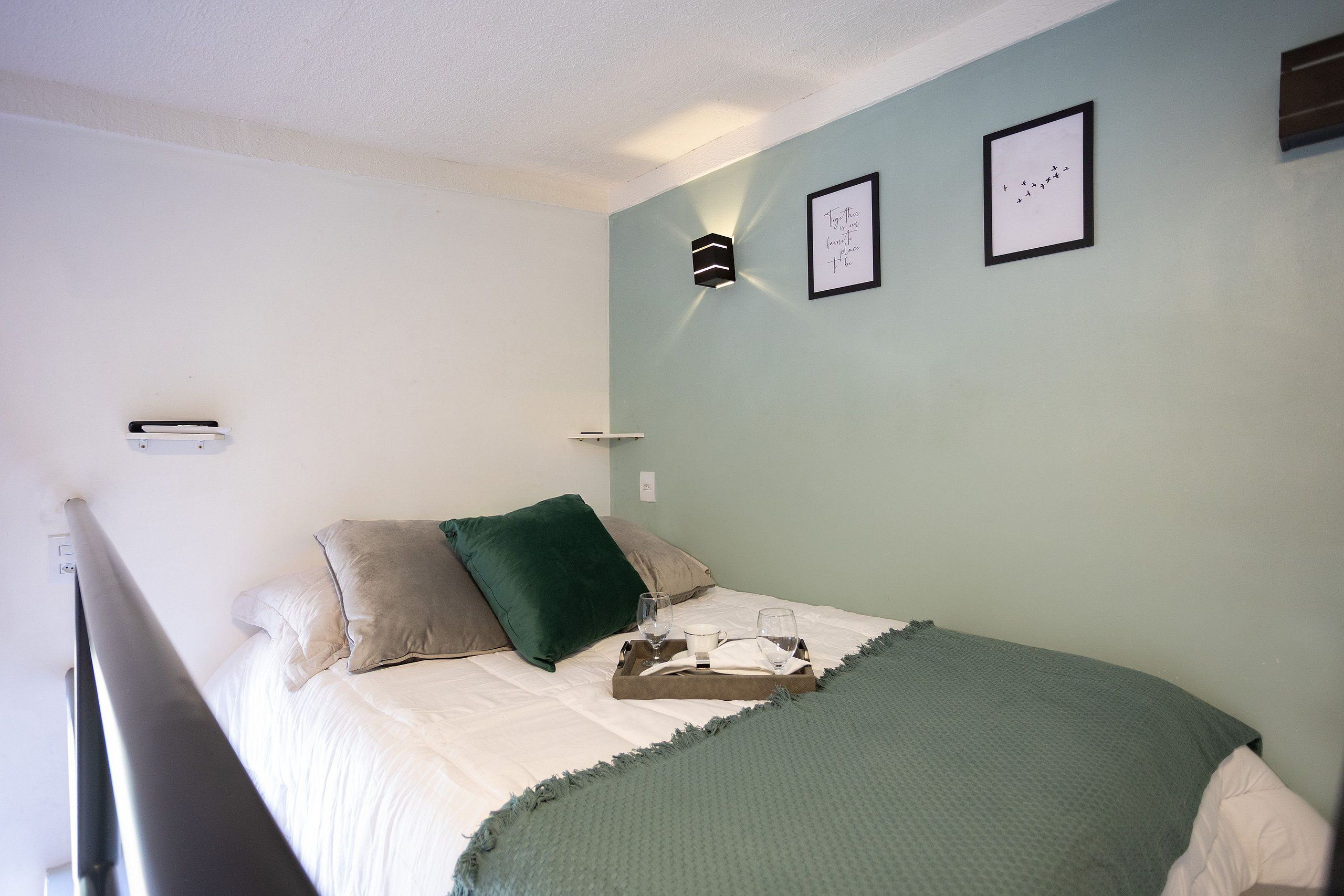 A bedroom with a bed, pillows, a bedside table with a glass, and a wall-mounted light fixture.