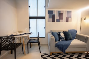 A modern living room with a sofa, a dining table with chairs, and a large window. - Residencial Laris - Lofts in Feira de Santana by Carpediem (Feira de Santana)
