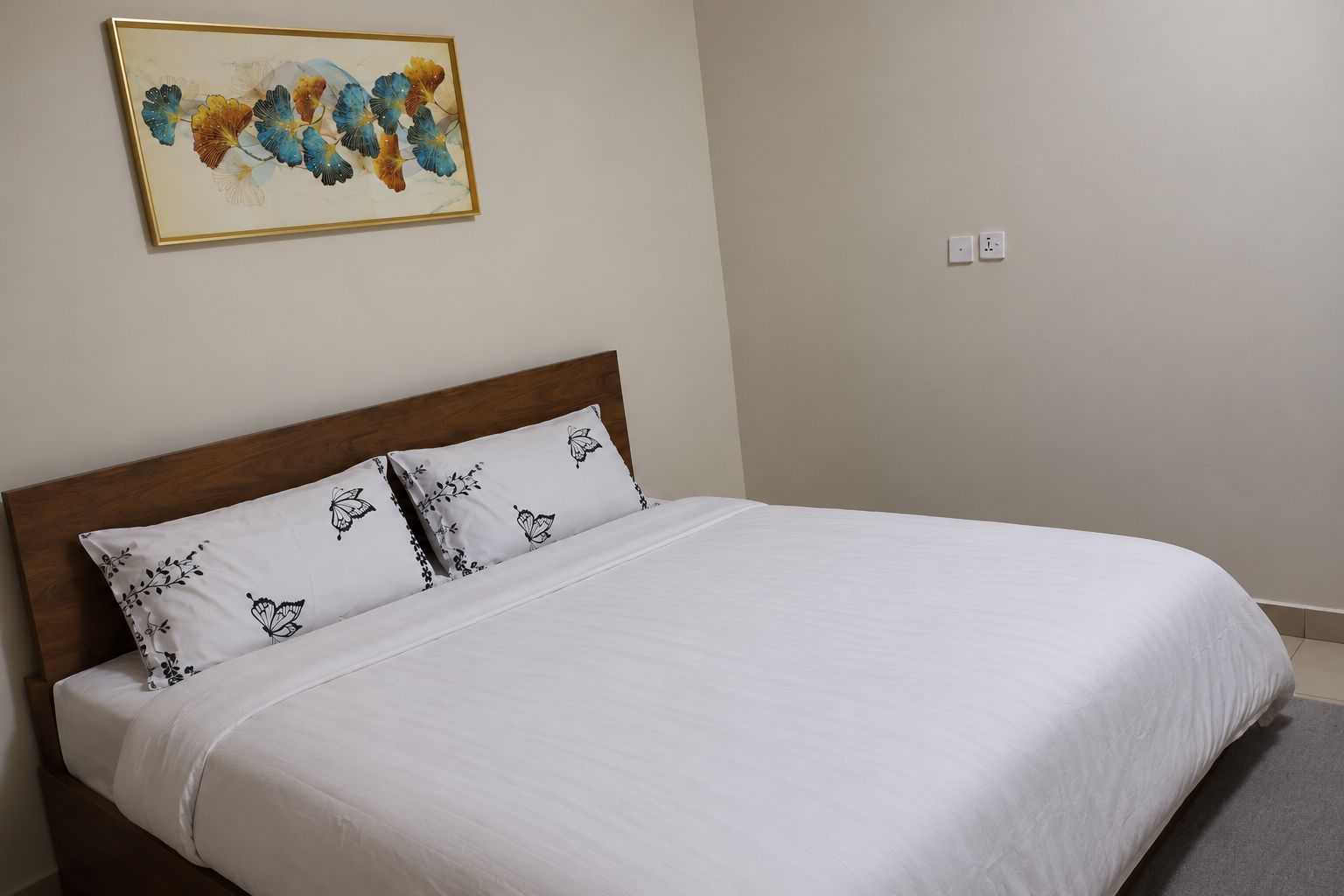 A neatly made bed with white bedding and patterned pillows, a wooden headboard, and a wall-mounted painting.