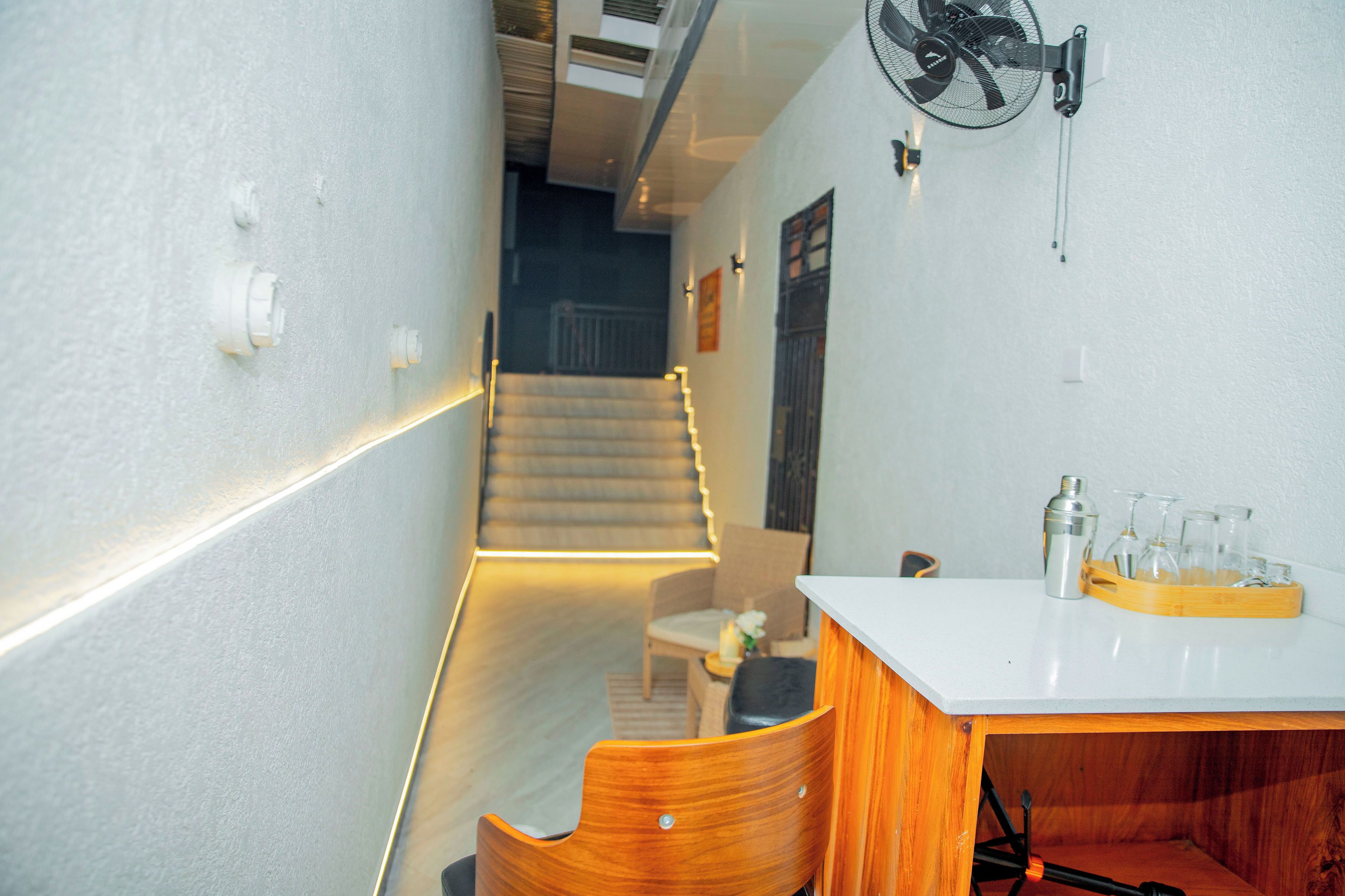 Un couloir étroit avec un escalier menant à un palier, un mur blanc orné d’un ventilateur mural et une chaise en bois.