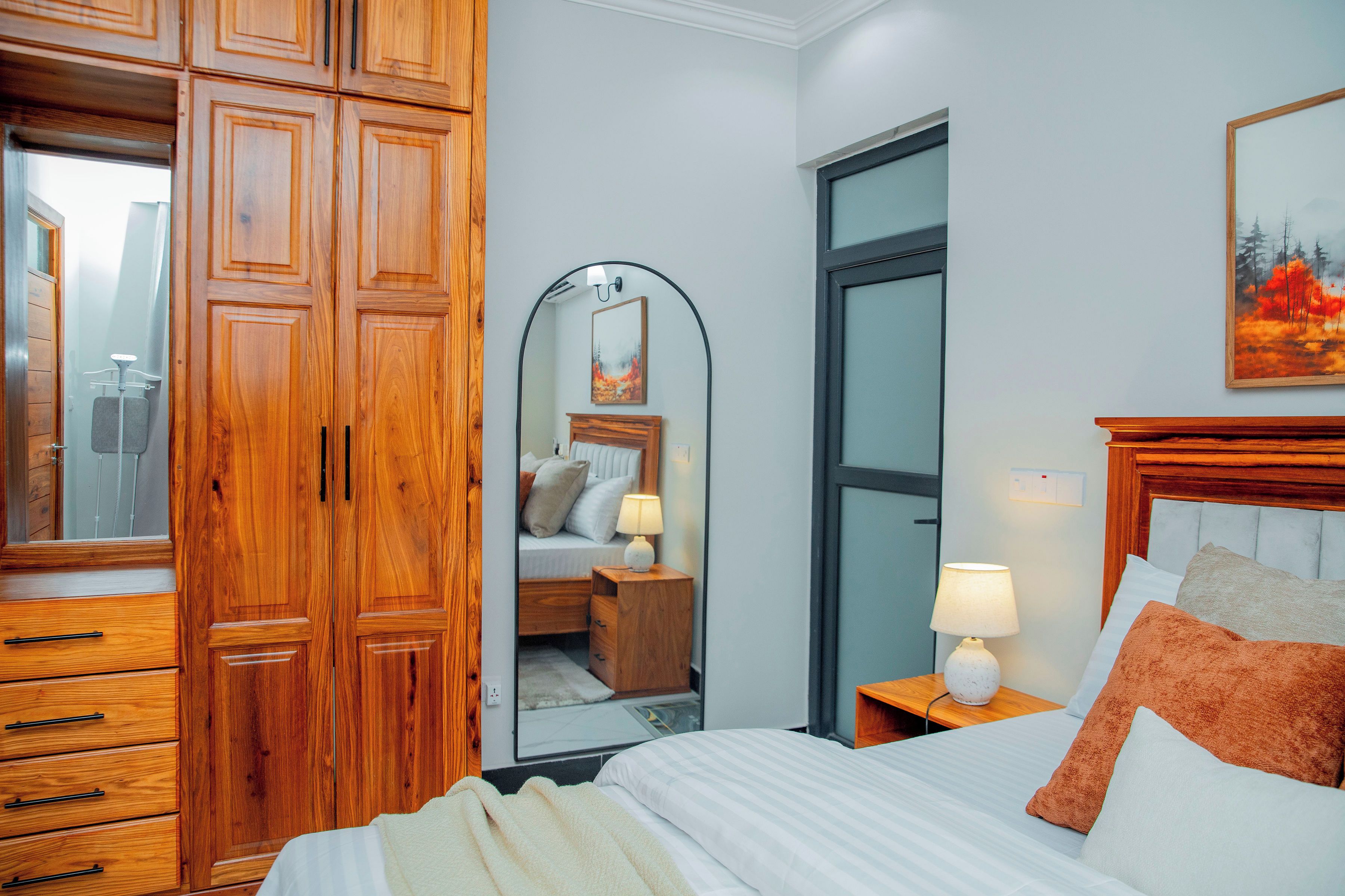 A hotel room with a wooden wardrobe, a bed with white bedding, a nightstand with a lamp, and a painting on the wall.