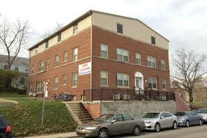 A multi-story brick building with a 'NOW LEASING' sign, a staircase, and several parked cars. - Steps from IU Campus & Jacobs School - 1BR Apartment (Bloomington)