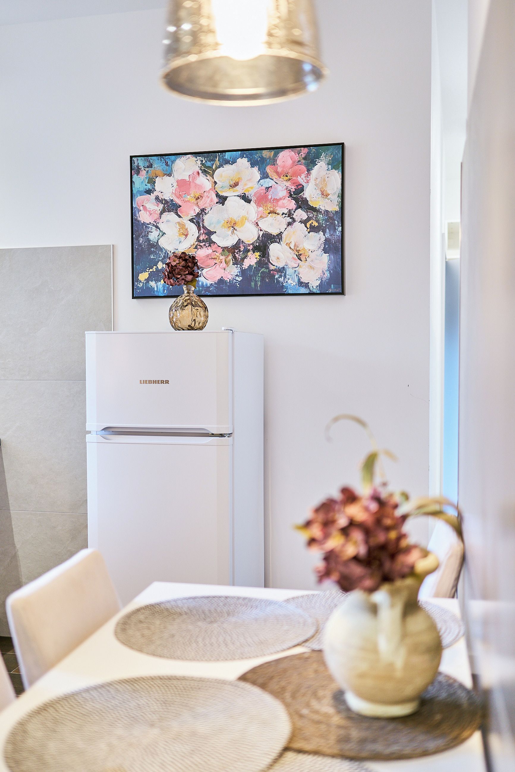 Una cocina moderna con un refrigerador blanco, una mesa de comedor con individuales y un florero con flores sobre la encimera.