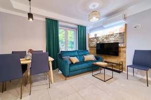 A modern living room with a teal sofa, a dining table with chairs, a coffee table, a flat-screen TV mounted on a wooden wall, and a ceiling fan. - Dom Solny (Polanica-Zdrój)