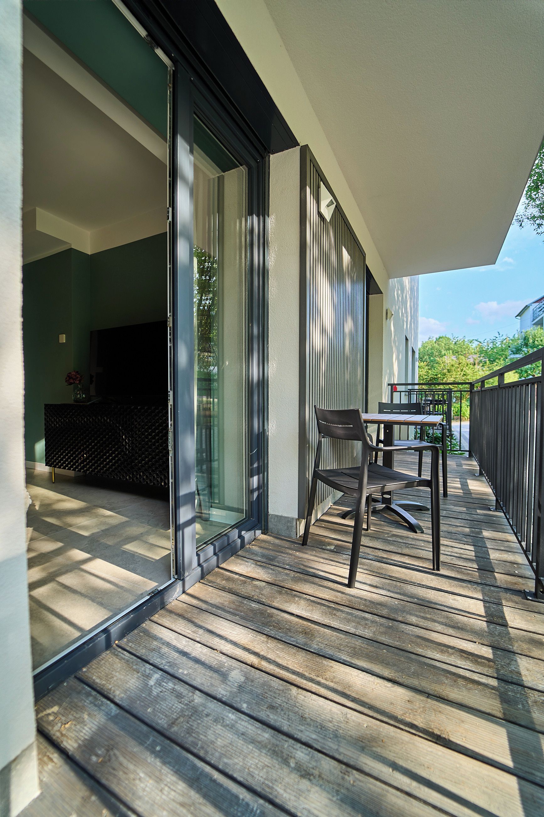 A modern outdoor deck with a dining table and chairs, a sliding glass door leading to a living area, and a wooden fence.