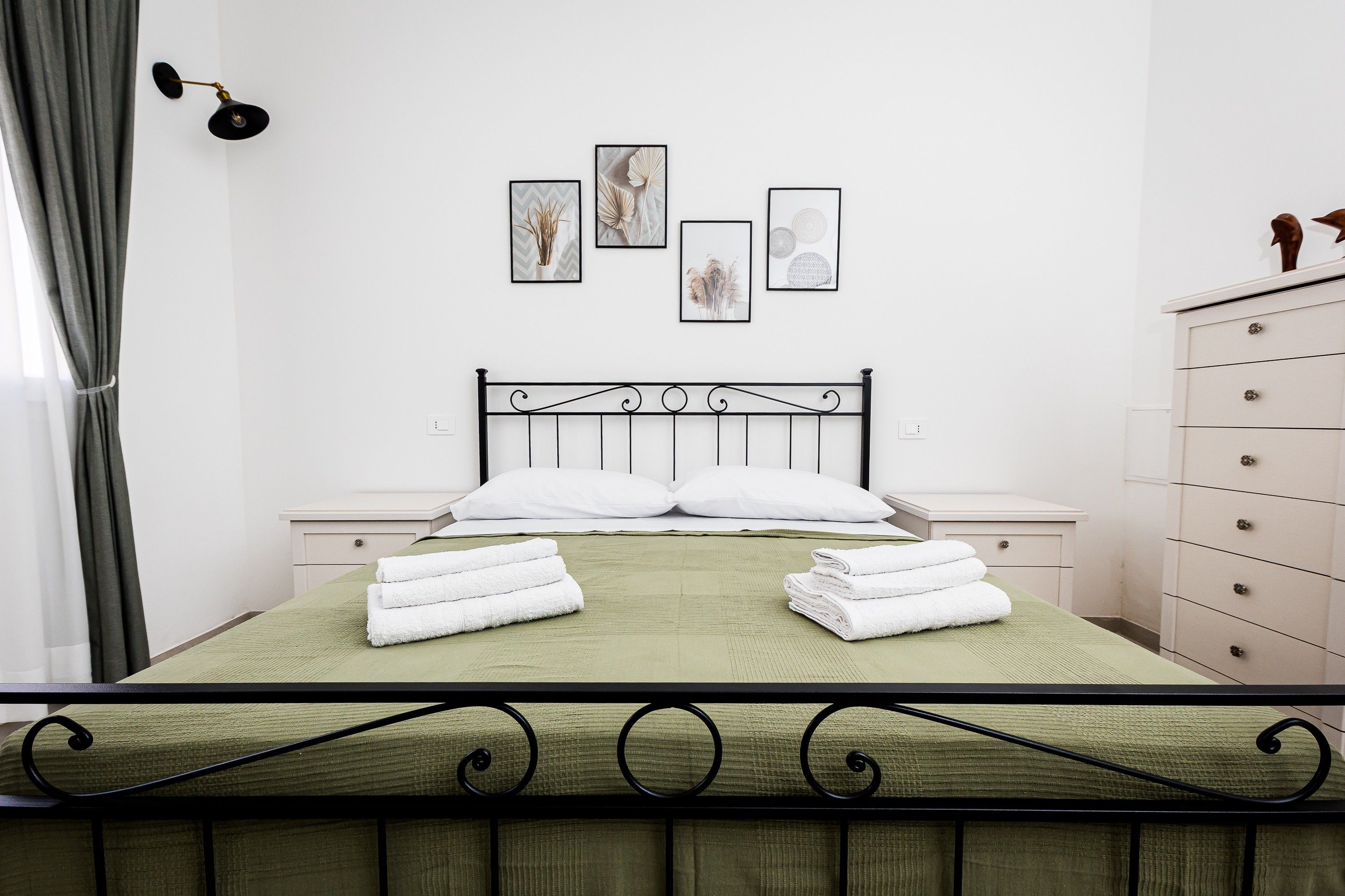 A neatly made bed with white pillows and folded towels, a bedside table, a dresser, and wall-mounted artwork.