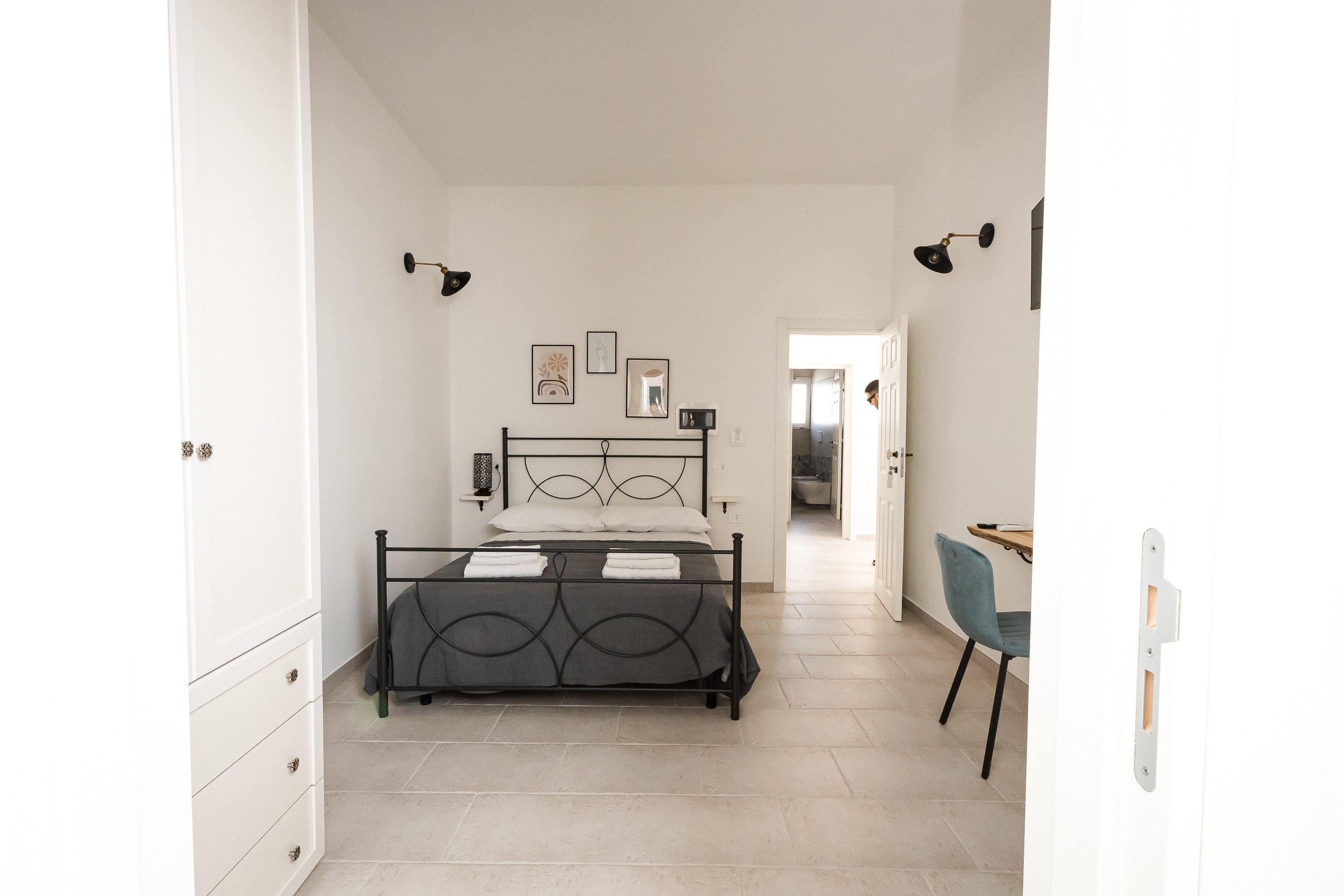 Un dormitorio con cama, escritorio y silla. Hay cuadros en la pared y una puerta que da a otra habitación.