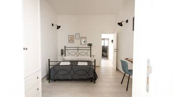 Un dormitorio con cama, escritorio y silla. Hay cuadros en la pared y una puerta que da a otra habitación.