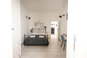Ein Schlafzimmer mit einem Bett, einem Schreibtisch und einem Stuhl. An den Wänden hängen Bilder, und eine Tür führt in einen weiteren Raum.