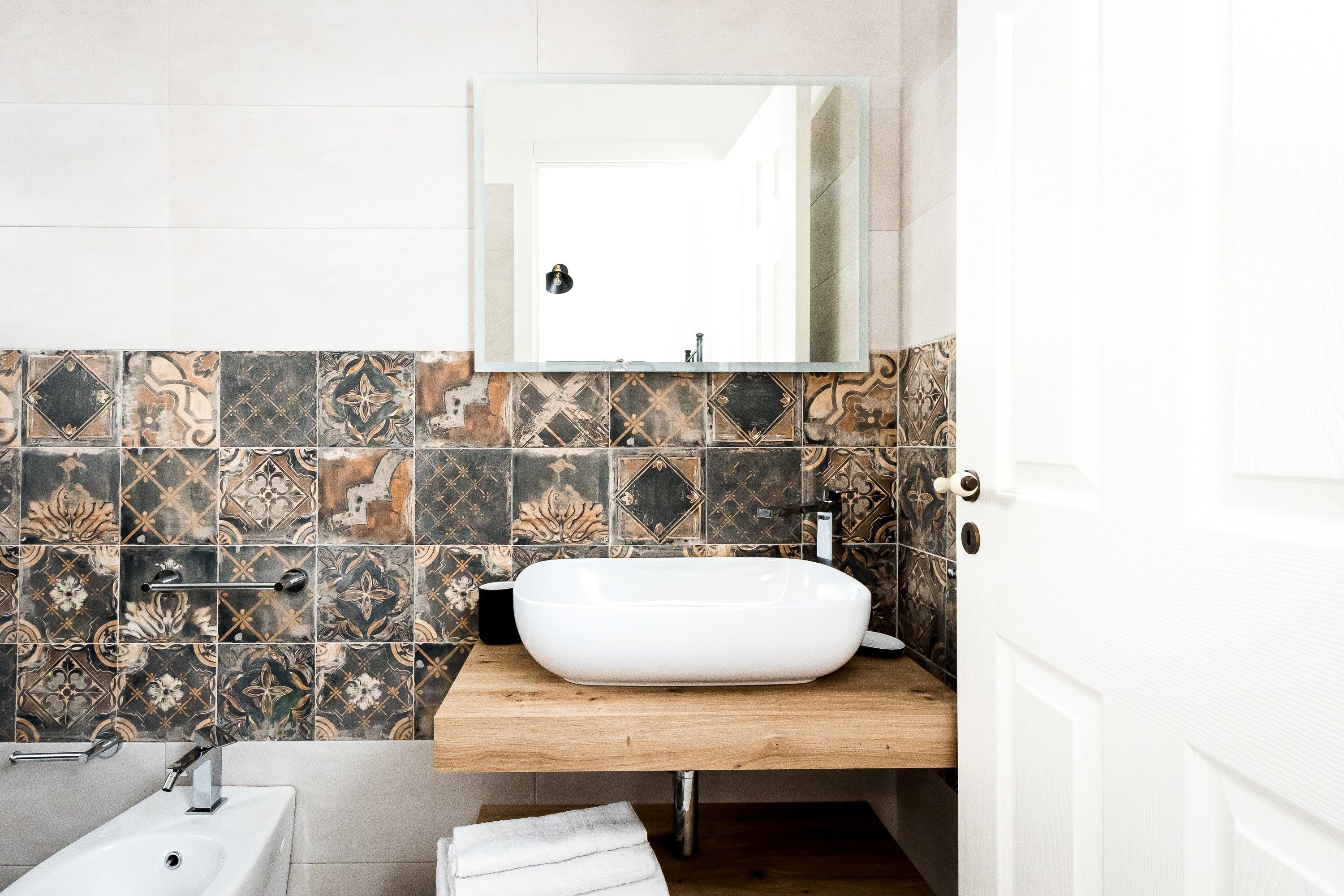Un bagno con un lavandino bianco, uno specchio sopra e una parete piastrellata con diversi motivi.