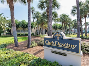 A sign for Club Destiny with a palm tree-lined driveway and a building in the background. - Destiny on the Cay | Executive Home, Private Pool + Golf Cart (Destin)