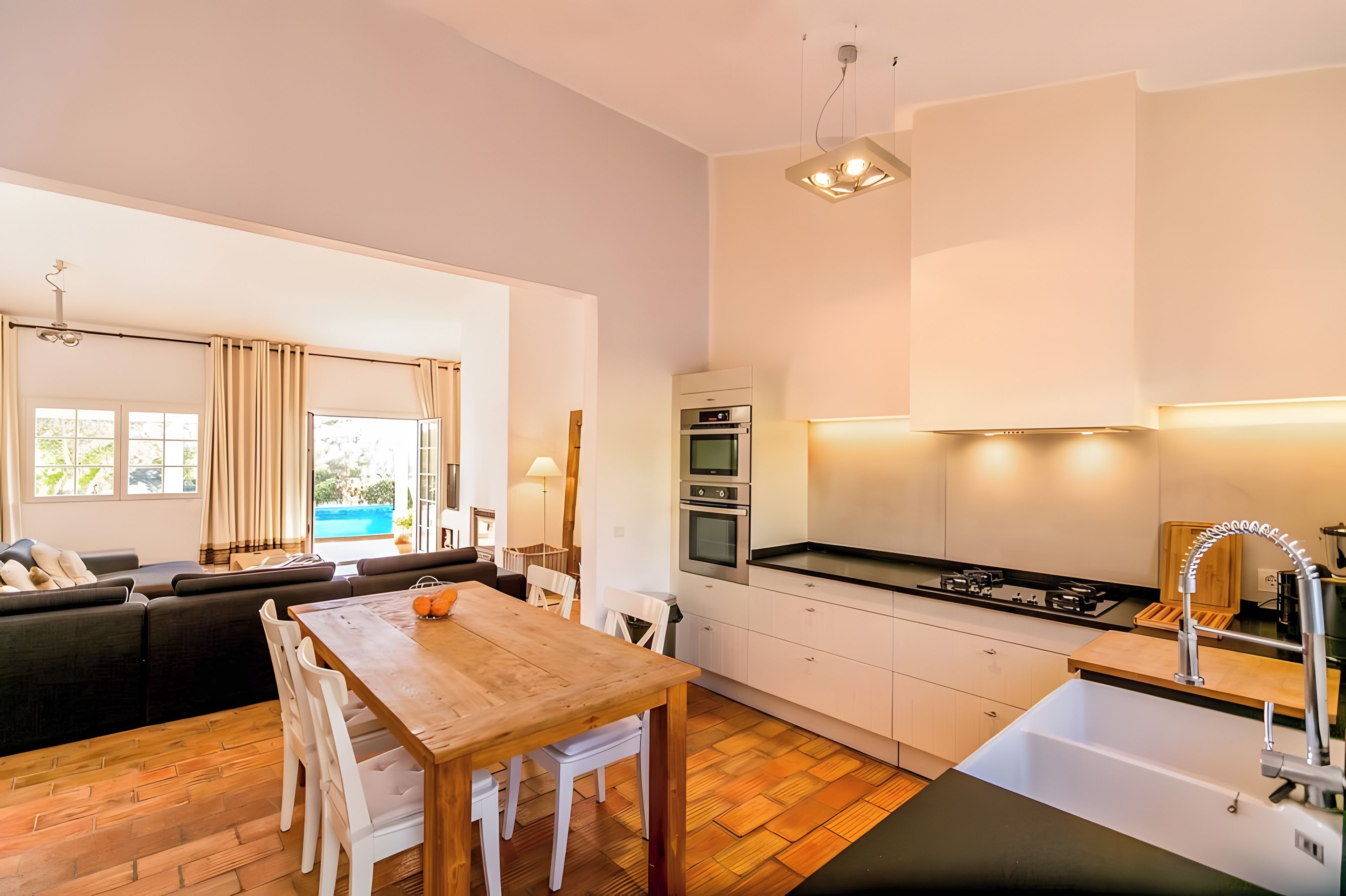 A modern kitchen with built-in appliances, a dining area with a wooden table and white chairs, and a view of a swimming pool through large windows.