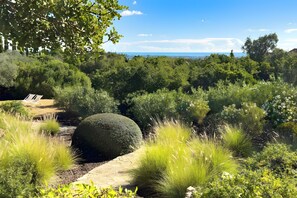 Een weelderige tuin met diverse planten, een bolvormig stenen element en een helder uitzicht op de zee op de achtergrond.