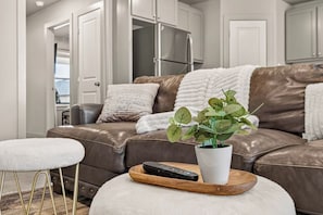 A modern living room with a leather sofa, a white ottoman, a potted plant, and a remote control on a wooden tray. - Beautiful Modern 3BR Home w/ Spacious Living Area (Buffalo Gap)